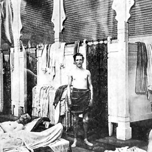 Shirtless white men in bath house with stalls and beds