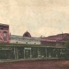 Horse drawn wagon on dirt street in front of brick storefront buildings with covered sidewalk