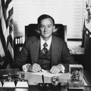 Portrait white man in suit and tie smiling at desk signing paper flanked by U.S. and Arkansas flags