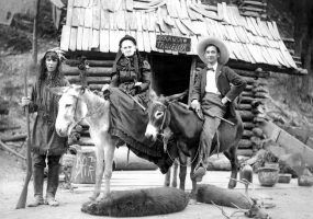 White man and old white woman on donkeys with white woman on foot dressed in Native American garb in front of log cabin with "Arkansaw Traveler" sign above the door