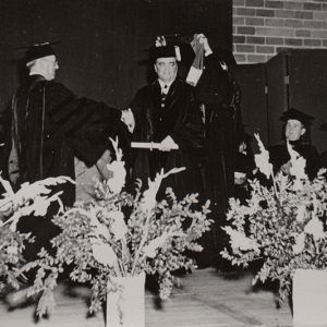 Graduation ceremony with young white man being hooded while shaking hands with older white man in robes on stage