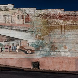 Faded mural depicting train depot building and service station with patrons and car on brick building