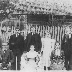 Family of eight with five standing and three sitting outside single-story house inside fence