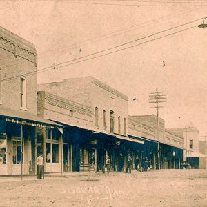 Men standing under awnings outside multistory brick storefront buildings on town street