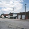 Street with brick storefronts and two-bay brick garage building with parked cars and two people standing