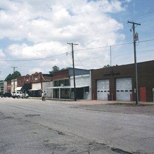 Street with brick storefronts and two-bay brick garage building with parked cars and two people standing