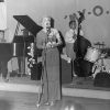 White man in dress and gloves singing with band on stage