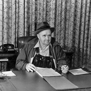 white man in overalls wind breaker and fedora sitting at desk with curtains behind him