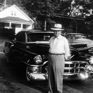 Older white man in hat standing in front of large shiny car in driveway