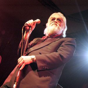 Older white man with beard at microphone on stage with younger white guitarist