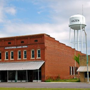 Two-story brick store building with awning and single-story storefront on street with water tower in the background