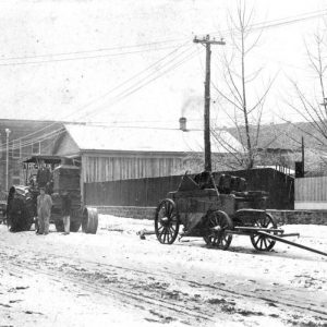 White men driving a tractor on a dirt road with buildings and telephone poles in the background