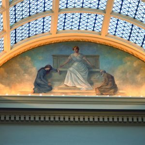 Glass roof with marble supports above a painting of a white woman in white robes with white man and woman bowing to her