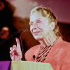 Older white woman with pulled back hair in a pink and black striped top and pink jacket talking at a lectern