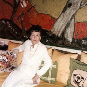 Older white woman in white sitting on couch with multicolored pillows and artwork on the wall behind her