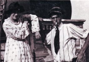 White man in shirt and tie smoking a cigarette with white woman in dress and hat