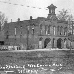 Multistory brick building with cupola alongside house on dirt street