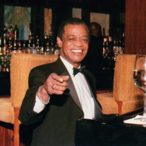 African American man in suit and bow tie sitting in bar pointing at camera