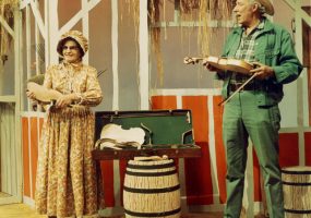 White man and woman folk costumes on stage with barn backdrop presenting violin making parts