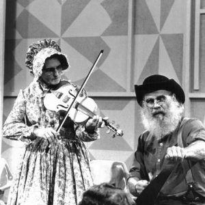 White man in suit smiling observing white woman and man playing violin and musical saw