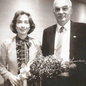 Young white woman holding flower bouquet standing next to older white man with glasses in suit and tie