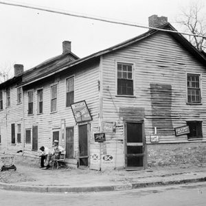 Run down two-story building on street corner with white men sitting in front parked cars on the street and billboard behind it