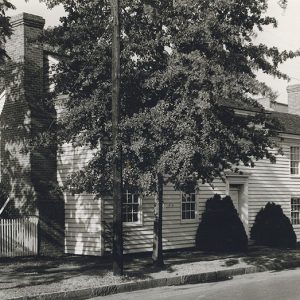 Two-story building with white siding and brick chimney behind tree on street corner