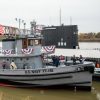 People standing on tugboat on river next to submarine with steel arch truss bridge in the background