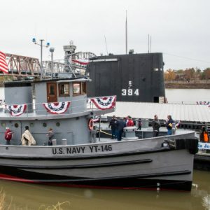 People standing on tugboat on river next to submarine with steel arch truss bridge in the background