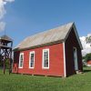 Bell tower next to single-story building painted red with open gable roof on grass