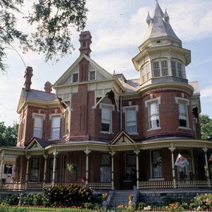 Ornate multistory brick house with covered wraparound porch with flags and a pointed tower