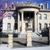 Three-story stone building with entrance covered with round covering and four columns behind black iron fence with three columns and a tree with purple flowers