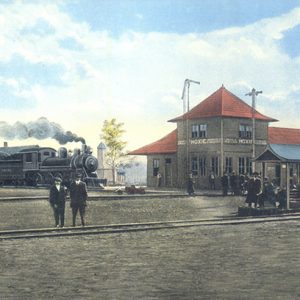 Steam train with cars arriving at multistory depot building with patrons in rail yard