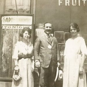 White man in suit and tie with two white women outside a storefront with "California Fruit" written on the window