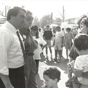 White man in shirt and tie alongside woman in sunglasses crowd talking to white woman holding a child amid an outdoor crowd