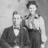 White man in suit and bow tie sitting with white woman in dress standing behind him