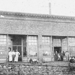 White workers and others standing outside brick building with stone wall and horse drawn wagon