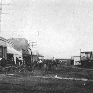 Men and horses on dirt road between rows of brick storefronts