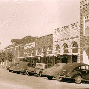 Parked cars sitting outside brick storefront buildings on town street with multistory buildings and house in the background