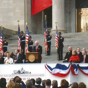 White man speaking at lectern on capitol steps with mixed crowd behind him