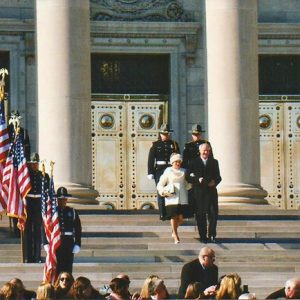 White man in suit and woman in white coat on capitol building steps with color guard policemen and crowd