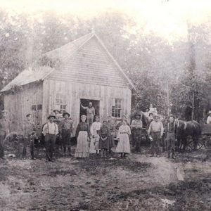 Group of white men women and children standing outside single-story building with A-frame roof and trees in background