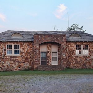 Single-story stone building with arched entrance way on grass with gravel driveway