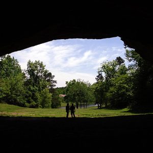 Two people standing at cave entrance with pond and building in the background as seen from inside the cave