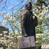 Statue of a man with suitcase in his right hand and hat over his heart on engraved pedestal next to tree with white flowers