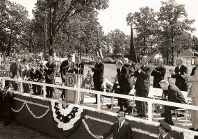 Decorated outdoor stage with white men and women clapping as a white man in suit approaches the lectern