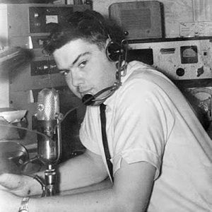 Young white man with headphones in radio booth with recording equipment around him