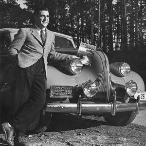 smiling dark-haired white man in tweed jacket dark pants and tie holding a book and standing in front of a shiny car