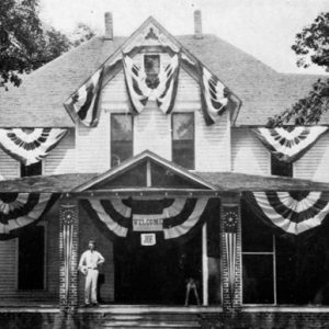 White man posing on front porch of multistory wood frame home that is covered in patriotic banners and a sign saying "Welcome Joe"