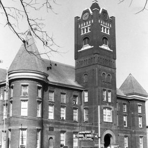 Multistory brick building with conical and pyramidal steeples and clock tower with robed figure sculpture on top